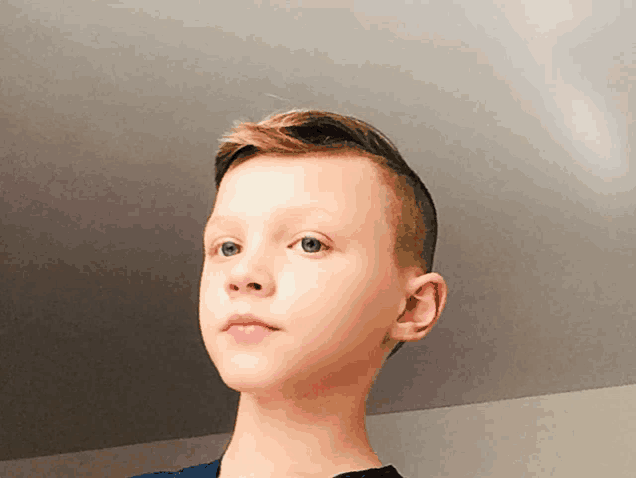 a young boy with a shaved head and a blue shirt looks at the camera