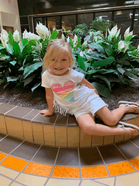 a little girl wearing a white shirt with a heart on it sits on a ledge