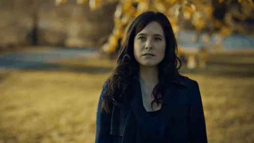 a woman in a blue coat stands in a field with a tree in the background