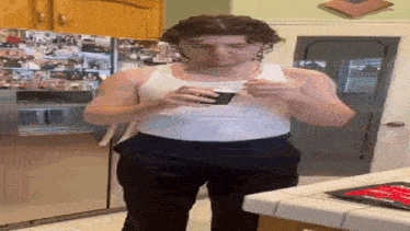 a man in a white tank top is drinking a cup of coffee in a kitchen .