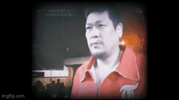 a man in a red jacket is standing in front of a crowd of people on a television screen .