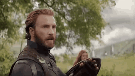 captain america is holding a shield in a field with a woman behind him .