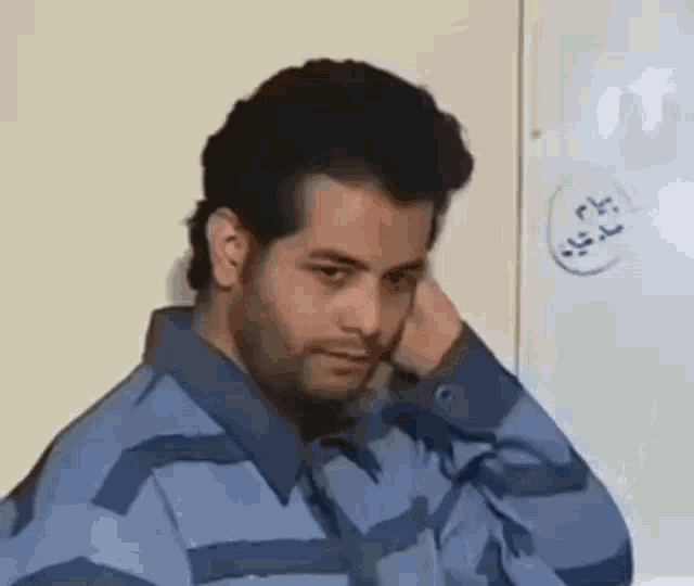 a man in a blue jacket is sitting in front of a white board and covering his face with his hand .