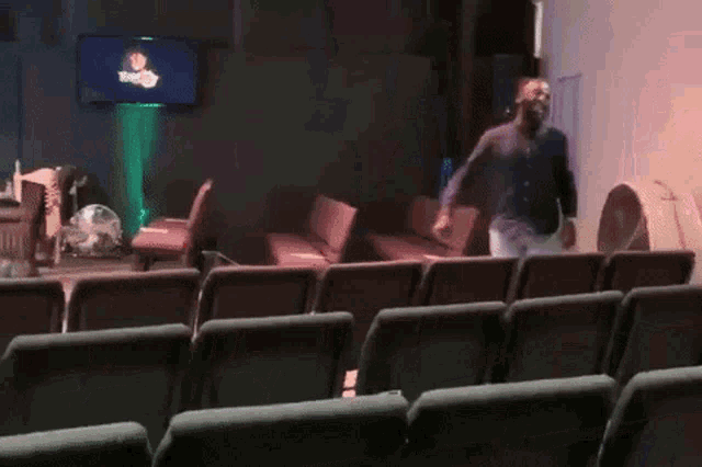 a man is dancing in an auditorium with a tv on the wall that says news