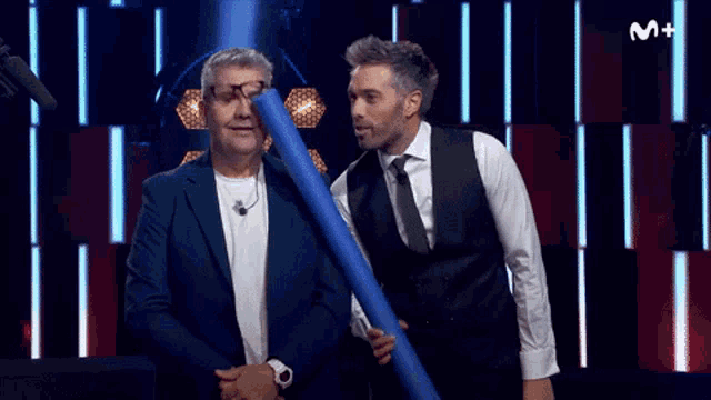 two men are standing next to each other and one is holding a blue tube with the letter m on the bottom right