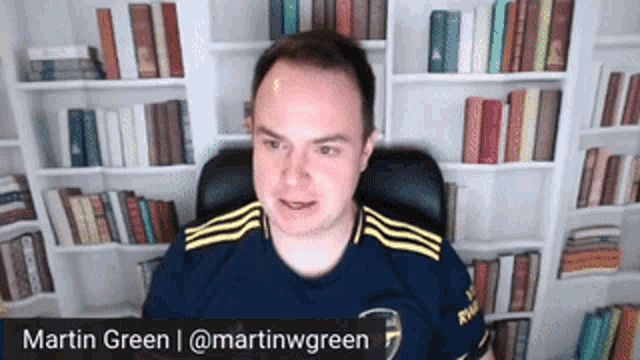a man in a blue shirt is sitting in front of a bookshelf and says martin green