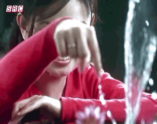 a woman in a red shirt is covering her face with her hand in front of a fountain with src written on the bottom right