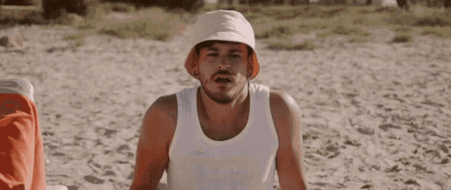a man wearing a white tank top and a white hat is sitting on the beach .