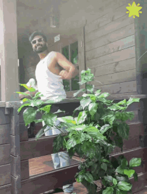 a man in a white tank top stands on a porch next to a plant