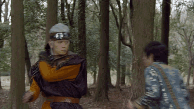 a man wearing a helmet and goggles stands in the woods with another man