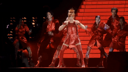 a woman in a red dress with a crown on her head is standing on a stage with a group of dancers .