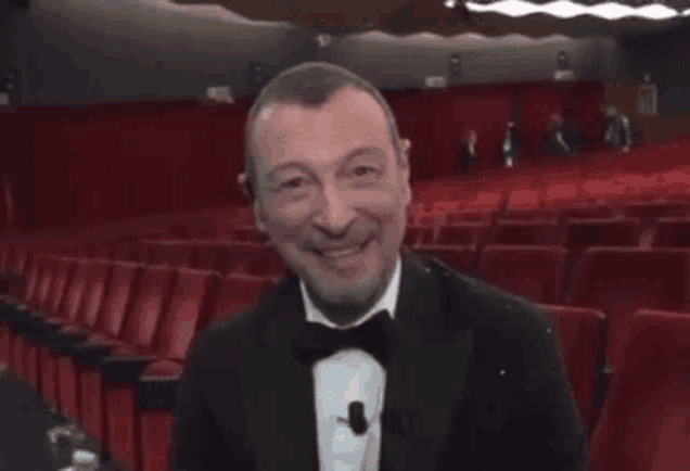 a man in a tuxedo and bow tie is smiling in an auditorium with red seats .