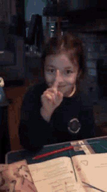a little girl is sitting at a desk making a shhh sign .