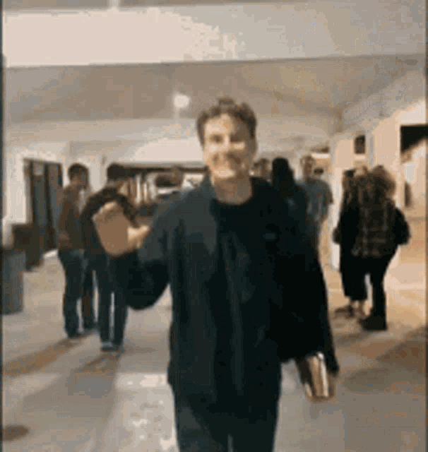 a man in a black jacket is walking down a hallway while holding a book .