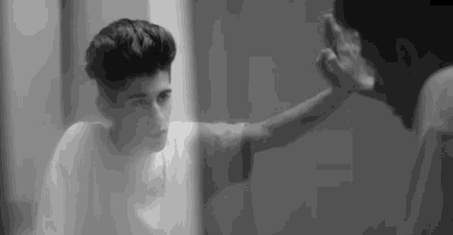 a black and white photo of a young man looking at himself in the mirror .