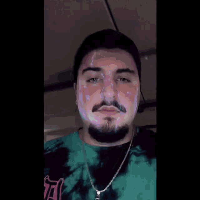 a man with a beard and a tie dye shirt is wearing a necklace and looking at the camera .