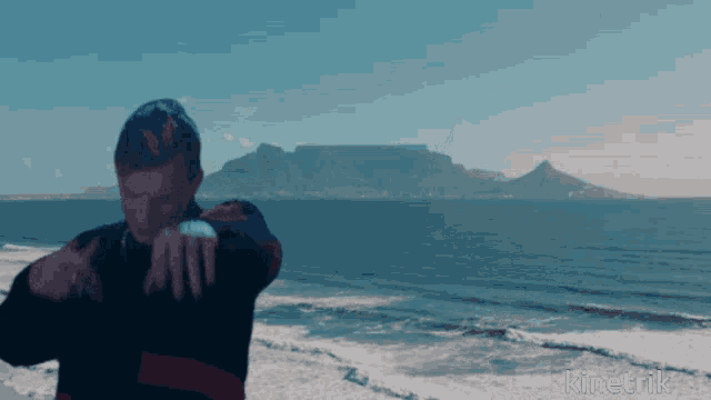 a man stands in front of a body of water with a mountain in the background and the word kinetik visible