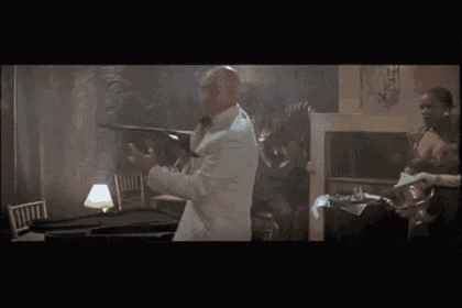 a man in a tuxedo is holding a gun in a room with a woman behind him