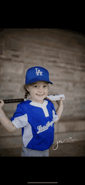 a little girl is wearing a los angeles dodgers jersey and holding a baseball bat