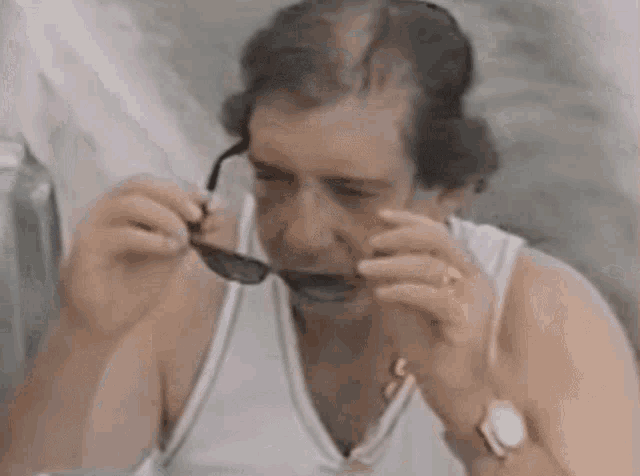 a man wearing a white tank top and a watch is eating something while holding a pair of sunglasses in his mouth .