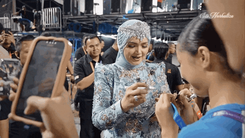 a woman in a hijab talks to another woman while holding a cell phone in front of a sign that says sin herbalife