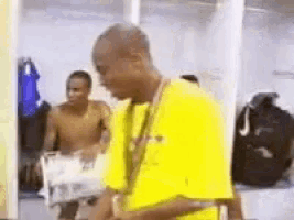 a man in a yellow shirt is standing in a dressing room with a shirtless man in the background .