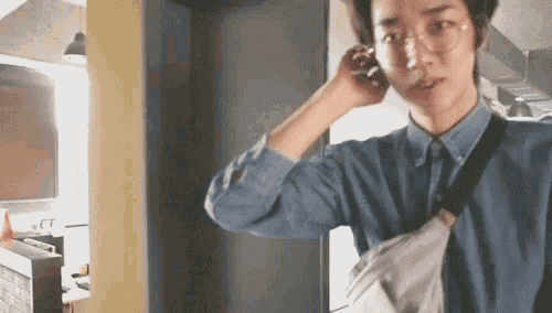 a young man wearing glasses and a denim shirt is talking on a cell phone .