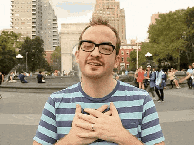 a man wearing glasses and a striped shirt holds his hands to his chest
