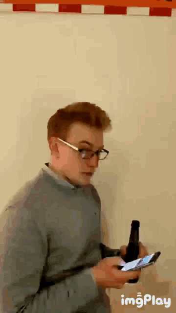 a man with glasses is holding a bottle of beer and looking at his phone .