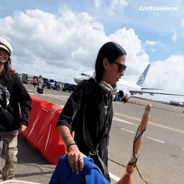 a woman wearing headphones walks on an airport runway with a plane in the background