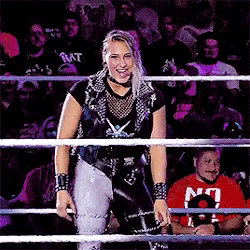 a woman in a wrestling ring with a crowd behind her and a man in a red shirt with the letter n on it