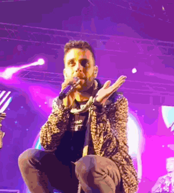 a man in a leopard print jacket is singing into a microphone on a stage with purple lights behind him .