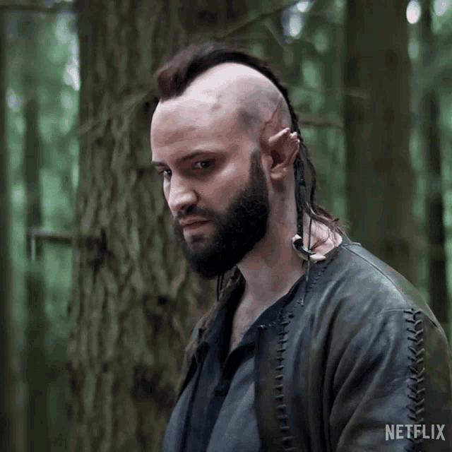 a man with a beard and mohawk is standing in the woods with a netflix logo on the bottom of his jacket