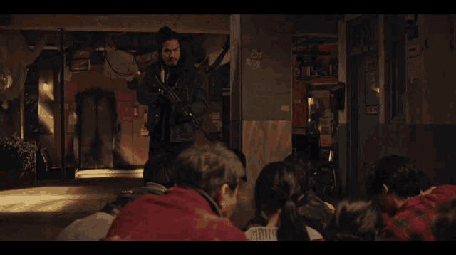 a group of people are gathered in a dark room with a man in a red jacket standing in the middle