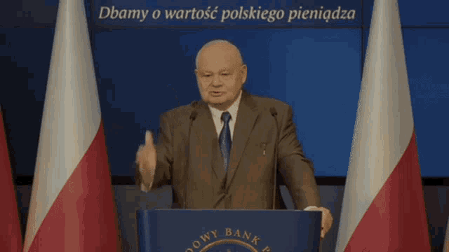 a man in a suit and tie stands at a podium giving a speech in front of two flags