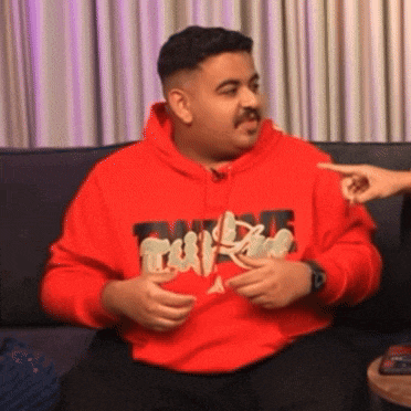 a man in a red hoodie is sitting on a couch talking to someone .
