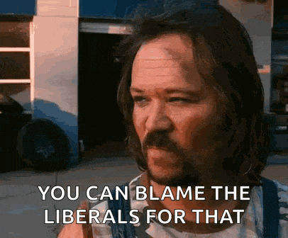 a man with long hair and a beard is standing in front of a garage and says `` you can blame the liberals for that ''