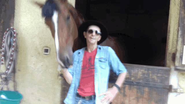 a man wearing a cowboy hat and sunglasses stands next to a horse in a stable