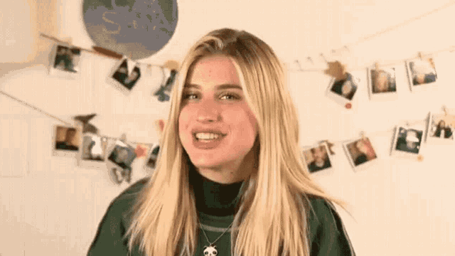 a blonde woman wearing a green sweater and a necklace is smiling in front of a wall with pictures hanging on it .