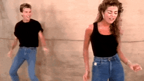 a man and a woman are dancing together in front of a wall . the woman is wearing high waisted jeans .