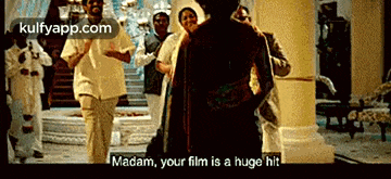 a group of people are standing in a room and one of them is saying madam your film is a huge hit .
