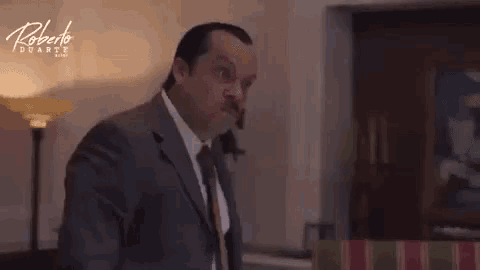 a man in a suit and tie is standing in a living room looking at the camera .