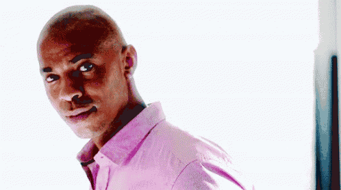 a man in a pink shirt is standing in front of a window and looking at the camera .