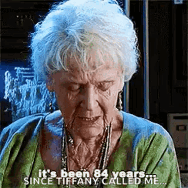 an elderly woman with gray hair is wearing a green sweater and necklace and is talking .