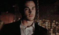a man in a suit and white shirt is standing in a dark room with lights behind him .
