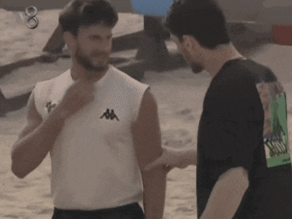 two men are standing next to each other on a beach . one of the men is wearing a white kappa shirt .