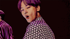 a man with purple hair is standing in front of a microphone and making a middle finger sign .