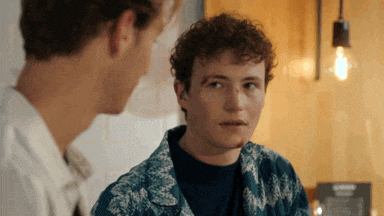 a man with curly hair looks at another man in a blue shirt