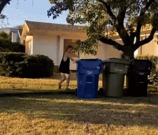a woman in a black dress is pushing a blue garbage can that says " recycle " on it
