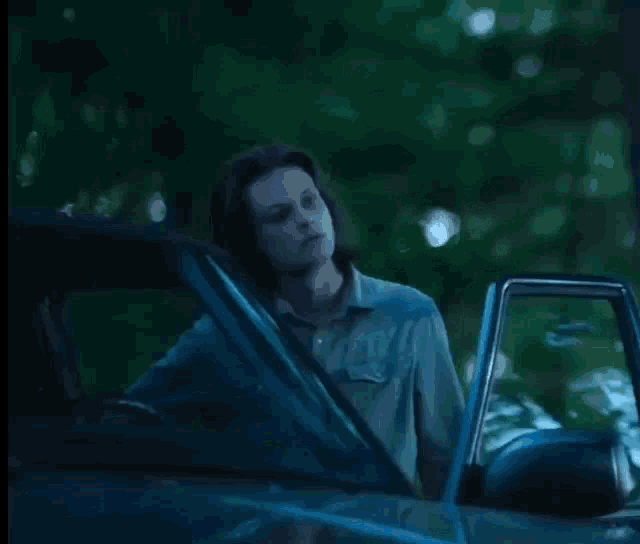 a man with long hair is standing next to a car in the dark .